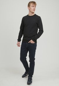 Fekete pulóver lekerekített nyakkivágással és bordás mandzsettával, sötétkék farmernadrággal és fekete sportcipővel párosítva. Egyszerű, letisztult dizájn.