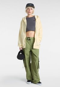 Světlě žlutá mikina s kapucí, šedý cropped top, zelené cargo kalhoty s kapsami, černá kulatá taška a černé tenisky. Krátké, světlé vlasy.