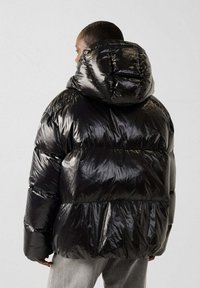 Chaqueta de pluma negra con acabado brillante, que cuenta con una gran capucha y secciones acolchadas horizontales. Costuras visibles y puños elásticos.