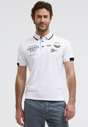 Mann trägt weißes kurzärmeliges Poloshirt mit segelthemenbezogenen Aufnähern und marineblauem Besatz, kombiniert mit blauen Shorts, vor schlichter Hintergrund.