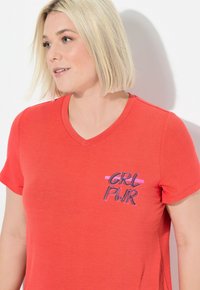 Punainen lyhythihainen V-pääntie t-paita, joka on valmistettu pehmeästä kankaasta ja jossa on kontrastiväreillä "GRL PWR" -grafiikka vasemmassa rintapielessä.