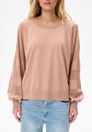 Kobieta ubrana w jasnoróżowy luźny sweter z siateczkowymi paskami na rękawach oraz jasnoniebieskie jeansy, stojąca na jednolitym tle.