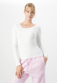 Blonde woman with bangs wearing a fitted white ribbed long-sleeve top and light pink drawstring pants, posing with one hand in pocket.