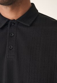 Polo noir texturé avec col, quatre boutons noirs et un motif à grille subtil.