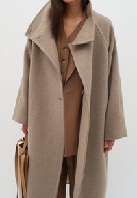 Cappotto in lana beige con un ampio colletto a punta, spalle basse e maniche lunghe. L'interno presenta un abbigliamento marrone complementare.