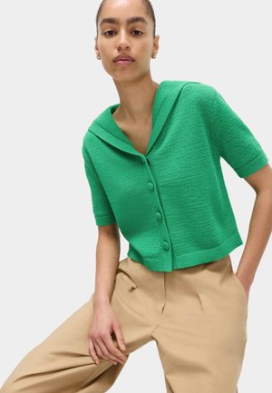 Frau trägt einen leuchtend grünen, kurzärmeligen Strickpullover mit Knöpfen und Kragen sowie beige, hoch taillierte Hosen und posiert mit einer Hand auf dem Knie.