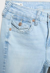 Gros plan sur un jean en denim bleu clair montrant la poche avant avec petite poche à bouton, les passants de ceinture et l'étiquette de taille 36 à l'intérieur de la ceinture.