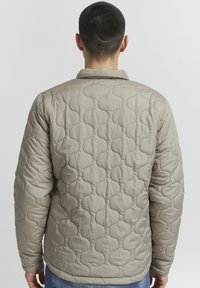 Beige quiltet jakke med et mønster, med krage, lange ermer og åpen rygg, laget av et lett stoff.