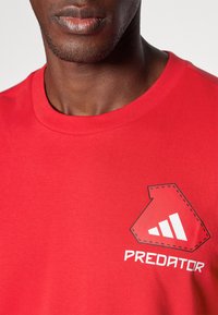 Camiseta de algodón roja con cuello redondo, textura suave y un gran gráfico blanco con la palabra "PREDATOR" en la parte delantera en costura negra contrastante.