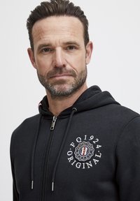 Sweat à capuche noir avec une fermeture éclair à l'avant, présentant un emblème circulaire avec le texte "EQ 1924 ORIGINAL". Tissu lisse et détails de capuche avec cordon.
