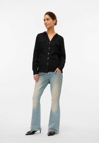 Schwarzes Hemd mit Knöpfen und zwei Taschen, kombiniert mit hellblauen Schlagjeans. Das Outfit wird durch schwarze hohe Schuhe ergänzt.