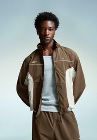 Brown and cream color-blocked jacket with a high collar, zipper front, and elastic cuffs, paired with a light grey tank top and shorts.