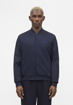 Rövid szőke hajú férfi, aki tengerészkék gombos zakót és hozzá illő sötét nadrágot visel, tiszta világos háttér előtt áll.