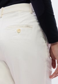 Gros plan sur une personne portant un pantalon de couleur crème avec une poche arrière boutonnée et une chemise noire à manches longues, main détendue sur le côté.