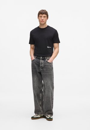 Junger Mann steht mit Händen in den Taschen, trägt ein schwarzes T-Shirt, lockere ausgewaschene graue Jeans und weiße und grüne Sneaker.