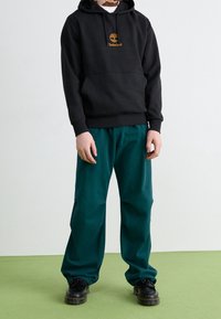Sudadera negra con capucha y logo de Timberland, combinada con pantalones sueltos de color teal y zapatos negros con cordones sobre un suelo verde.