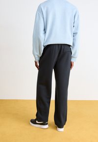 Ljusblå oversized sweatshirt, svarta sweatpants och svarta sneakers med vita detaljer, sedd bakifrån mot ett gult golv.