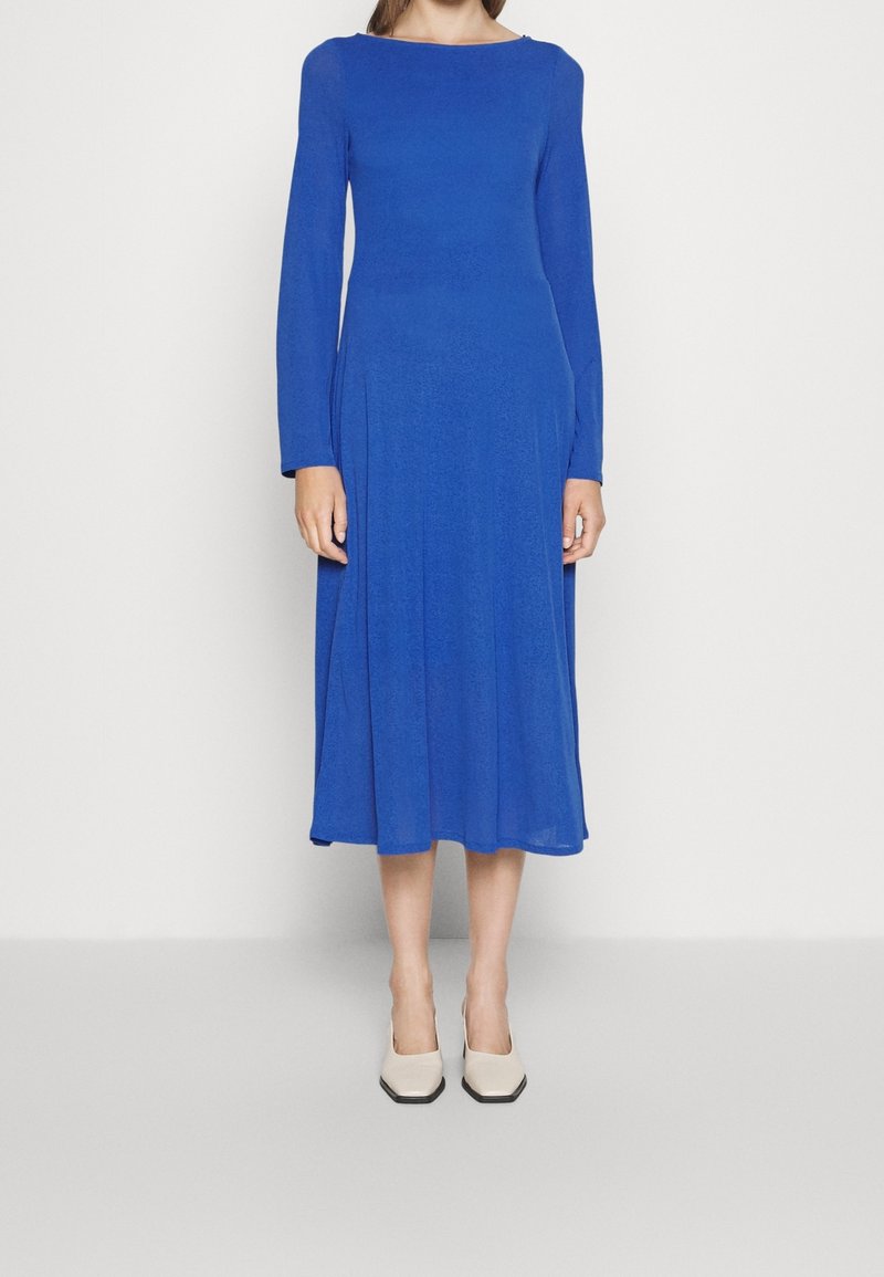 Femme portant une robe bleu roi à manches longues, longueur midi, associée à des talons beiges à bout carré, debout devant un fond clair uni.