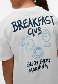 Camiseta blanca con el texto "BREAKFAST CLUB" y ilustraciones de un croissant, una taza de café y un postre en tonos azules. "DISFRUTA CADA MAÑANA" abajo.