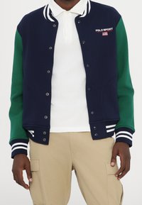 Marint och grönt collegejacka med vit randig krage och muddar, tryckknappar, broderad logotyp och avslappnad passform. Bärs över en vit pikétröja.