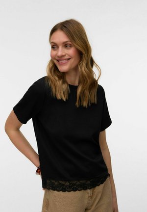 Femme aux cheveux ondulés mi-longs, portant un haut noir à manches courtes avec un ourlet en dentelle et un pantalon beige, souriante et regardant sur le côté.