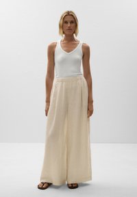 White ribbed tank top, paired with wide-leg, light beige pleated trousers. Feet in brown sandals. Neutral background.