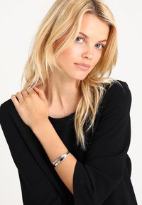 Brățară metalică cu o finisare argintie lustruită, având elemente de design geometric. Purta pe un încheietura mâinii, completând o bluză neagră.