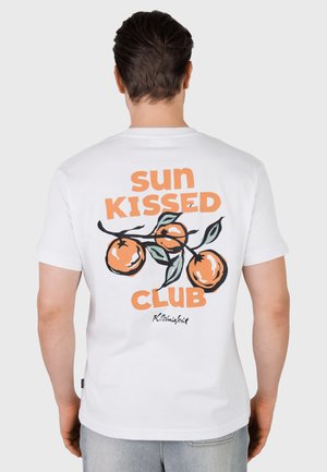 Man draagt een wit t-shirt met een grafiek van een sinaasappelboomtak en de tekst "sun kissed club" op de achterkant tegen een grijze achtergrond.