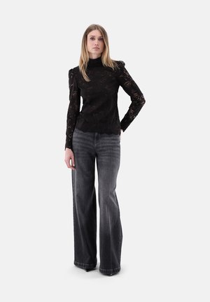 Femme debout portant un top en dentelle noire à manches longues et un jean noir délavé taille haute à jambes larges sur un fond clair uni.
