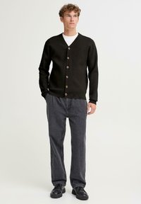 Tamsiai žalia megzta kardiganas su V formos iškirpte, šešiais mediniais mygtukais, ribinėmis rankogaliais ir apačia. Dėvimas virš baltos marškinės su pilkomis laisvo kirpimo kelnėmis.