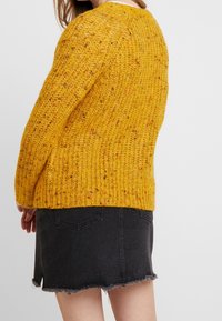 Maglione giallo lavorato a maglia con un motivo testurizzato, che presenta una vestibilità ampia e polsini a coste, abbinato a una gonna in denim nera con orlo sfrangiato.