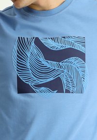 Camiseta de color azul claro con un gráfico cuadrado en navy oscuro que presenta patrones abstractos de hojas azules. Material de algodón con una textura suave.