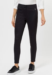 Pantaloni neri slim-fit con un motivo testurizzato, due cerniere frontali e una finitura liscia. Abbinati a sneakers nere e una maglietta bianca.