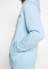 Felpa leggera blu con tasca frontale, cappuccio con cordoncino e logo ricamato. Presenta polsini e orlo a costine, realizzata in tessuto morbido.
