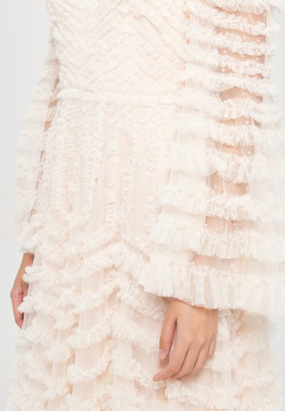 Close-up van een lichtroze jurk met gelaagde ruffles en doorzichtige mouwen op de arm en hand van een persoon.