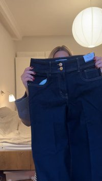 Person, der holder mørkeblå vide jeans med dobbelte guldknapper i et hyggeligt, blødt oplyst soveværelse.