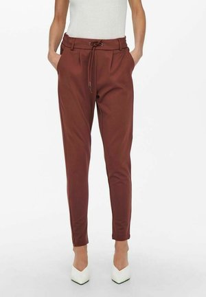 Frau trägt braune hoch taillierte, konisch geschnittene Hose mit Kordelzug und Taschen, kombiniert mit weißen spitzen Pumps und einem ärmellosen weißen Oberteil.