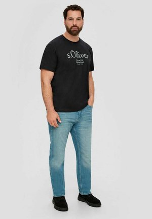 Man with dark hair and beard stands wearing black "s.Oliver" t-shirt, light blue jeans, and black shoes against a plain gray background.