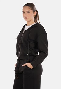 Felpa nera con zip, cappuccio regolabile, tasche frontali e polsini a costine. Realizzata in tessuto morbido con una vestibilità comoda e un design semplice.