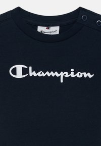 T-shirt en coton bleu marine avec un col rond, arborant un logo "Champion" blanc et des boutons-pression sur l'épaule pour un enfilage facile.