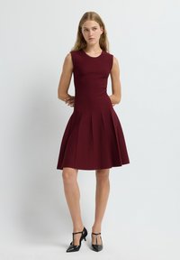 Robe sans manches bordeaux avec un corsage ajusté et une jupe évasée plissée. Texture lisse avec des détails de coutures visibles. Portée avec des chaussures à talons noires.