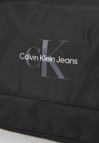 Borsa in nylon nera con logo bianco "Calvin Klein Jeans" e un design "CK" in grigio ben in evidenza. Texture liscia con dettagli di cucitura sottili.