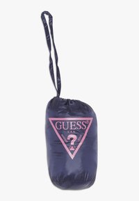 Navy blue drawstring bag made of shiny fabric, featuring a large pink triangular logo with "GUESS" and text "WASHED JEANS."