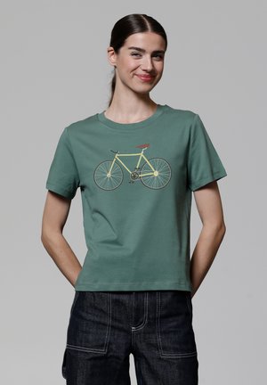 T-shirt en coton vert avec un graphique de bicyclette jaune et des accents marron, col ras du cou et manches courtes. Porté avec un pantalon en denim foncé.