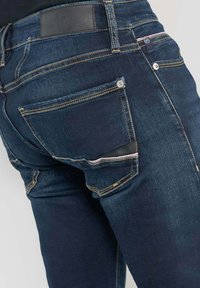 Jeans bleu foncé avec une texture lisse, des coutures jaunes et une patch en cuir au niveau de la taille. Possède une poche arrière avec un bouton.