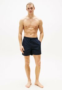 Marineblaue Schwimmshorts mit elastischem Bund, verstellbarem Kordelzug, Seitentaschen und leichtem Stoff. Das Modell steht barfuß und blickt nach vorne.