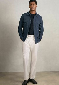 Marineblaues Hemd-Jackett mit zwei Brusttaschen, über einem schwarzen Hemd getragen. Kombiniert mit weißen maßgeschneiderten Hosen und schwarzen Mokassins. Glatte Textur.