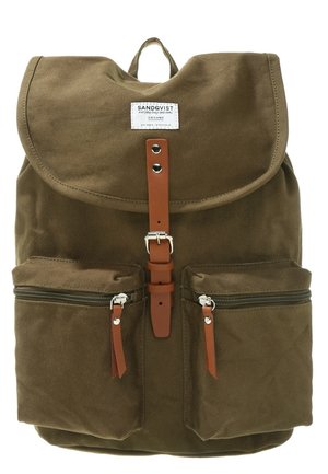 Brązowy płócienny plecak z klapą na górze, skórzanym paskiem na klamrę, dwoma przednimi kieszeniami na zamek oraz tekstylną etykietą z nazwą marki.