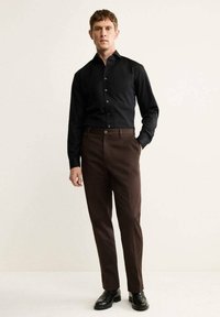 Maglietta nera a maniche lunghe con colletto alla mandarino abbinata a pantaloni marroni su misura, completata da scarpe nere. Design semplice, tessuto morbido.