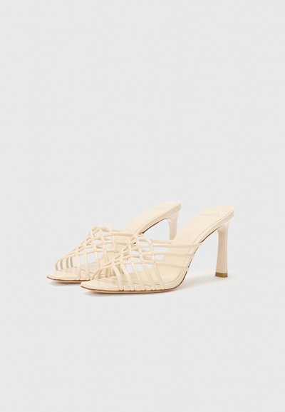 Mules de tacón alto color beige con diseño tejido y punta abierta, con finas tiras, sobre un fondo blanco liso.